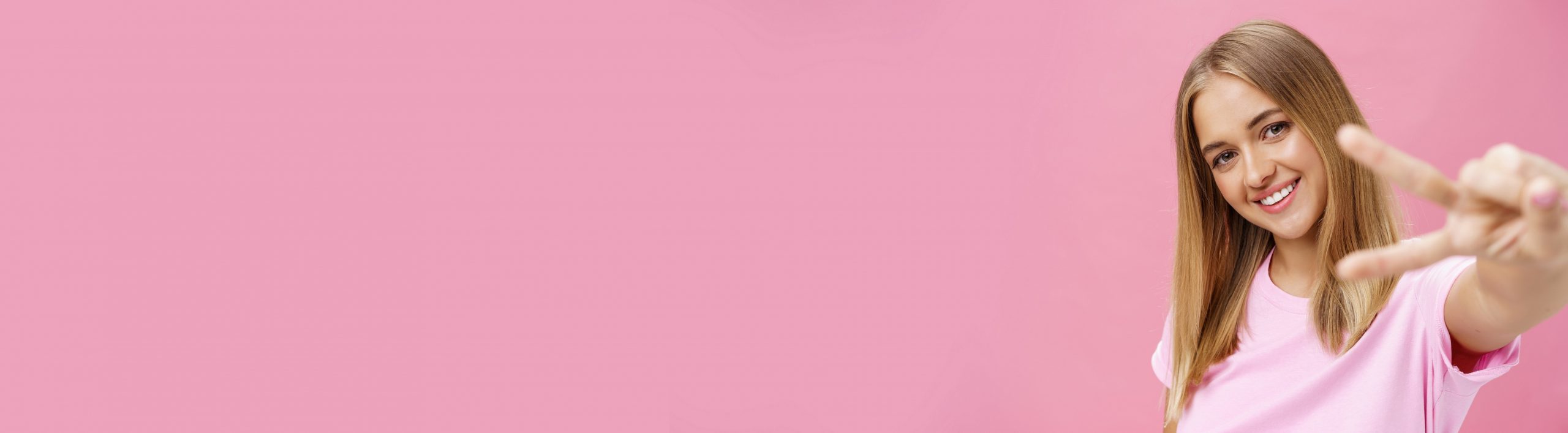 Peaceful and joyful good-looking young girl with straight blond hair in t-shirt pulling hand with peace sign towards camera, tilting head smiling friendly posing against pink background.