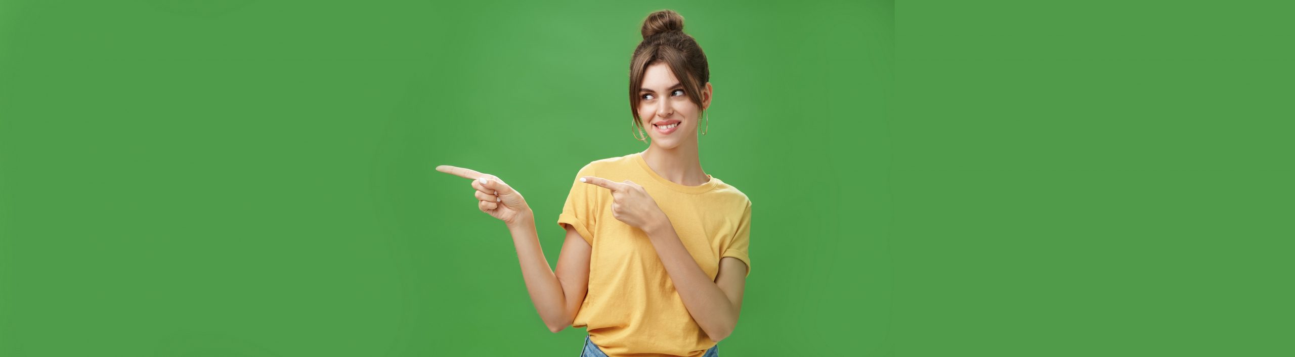 Waist-up shot of charismatic happy and carefree charming woman in yellow t-shirt pointing and looking left enthusiastic and pleased smiling cheerfully posing against green background delighted